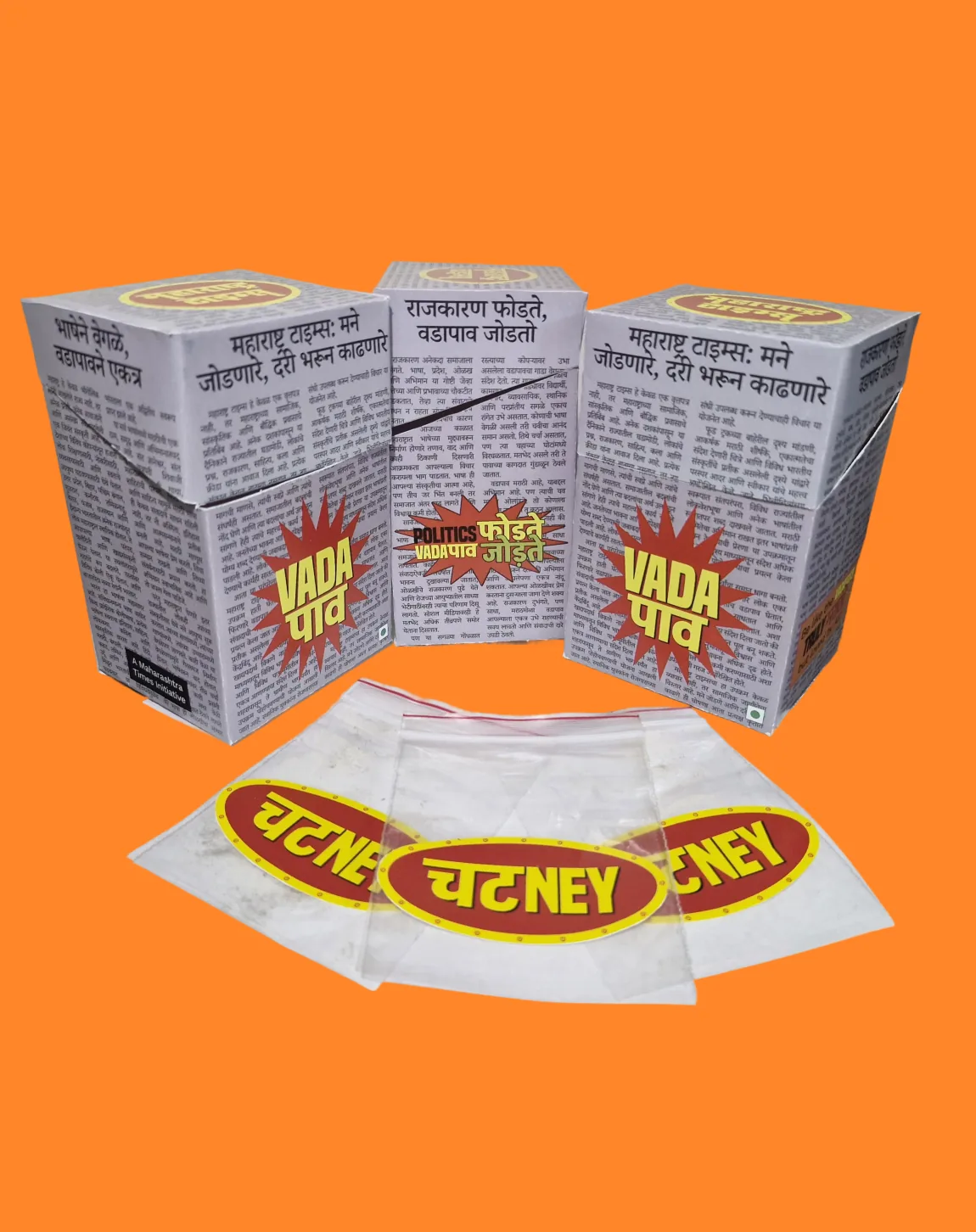 Three vada pav boxes on orange, chutney sachets fanned out in front of them.
