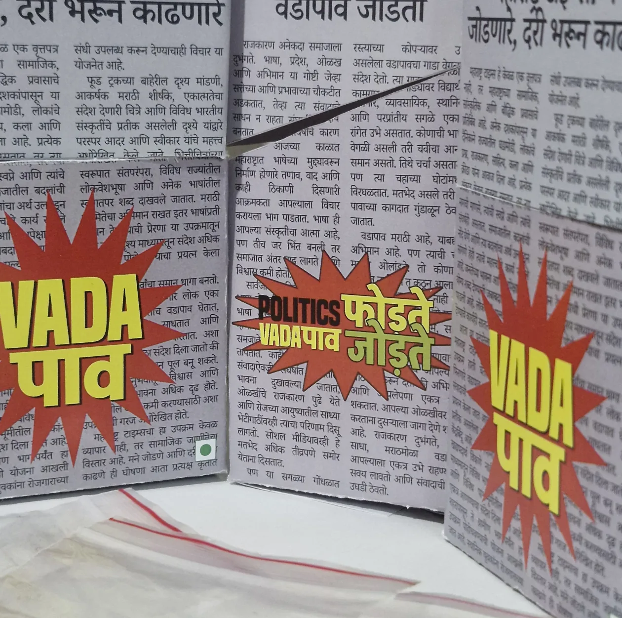 The vada pav box, printed as a Marathi newspaper, with “VADA पाव” starbursts on each face and a chutney sachet in front.