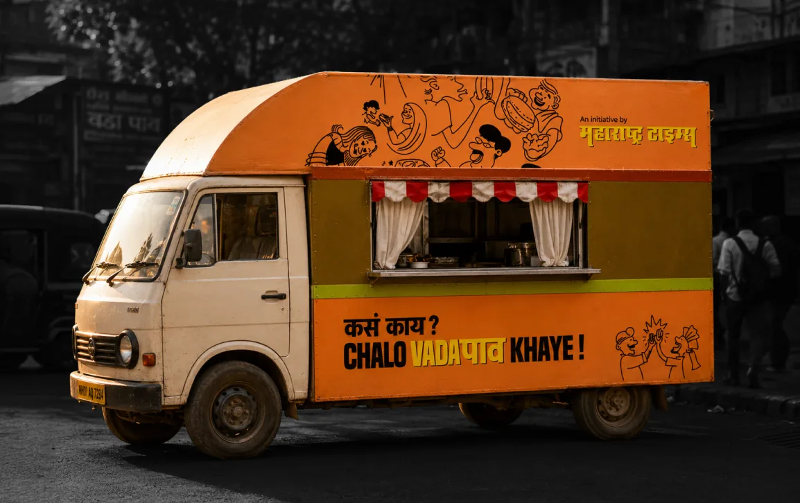 An orange food truck on a street: “Kasa kaay? Chalo vada pav khaye!”