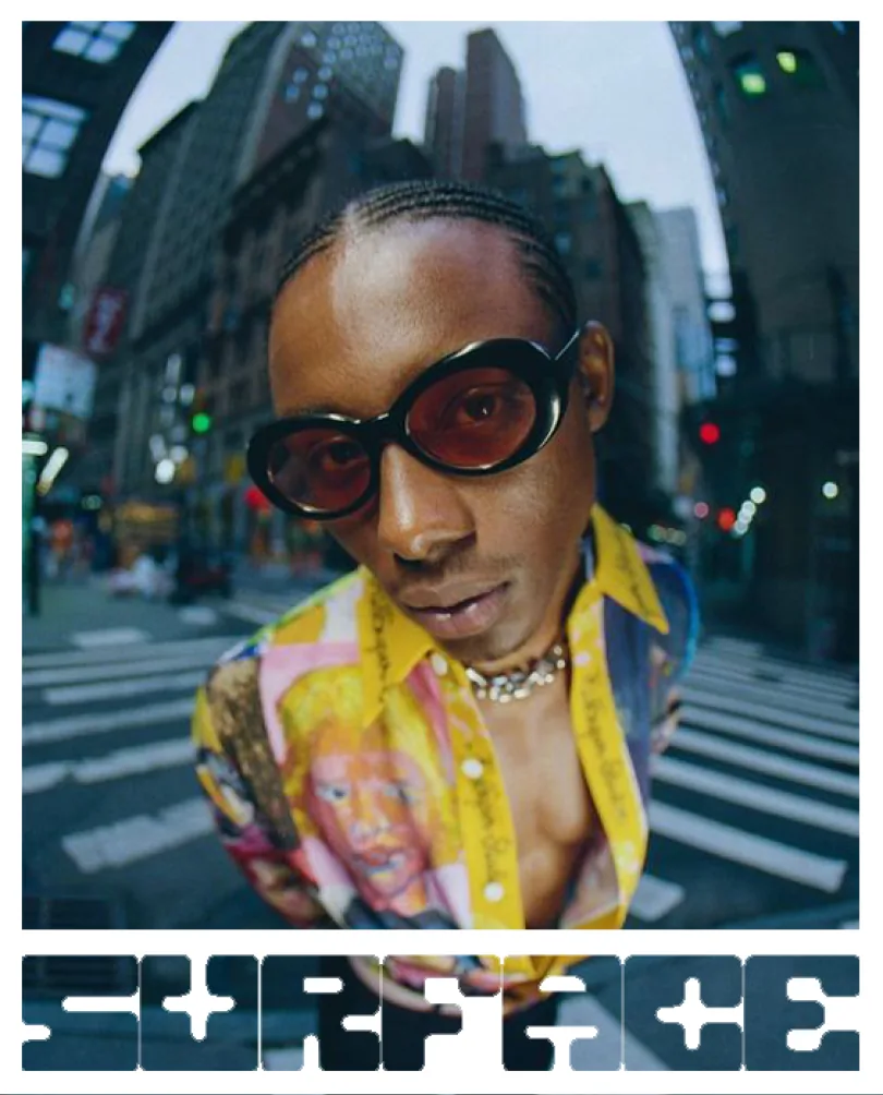 A model in a printed shirt and round sunglasses on a city crossing.