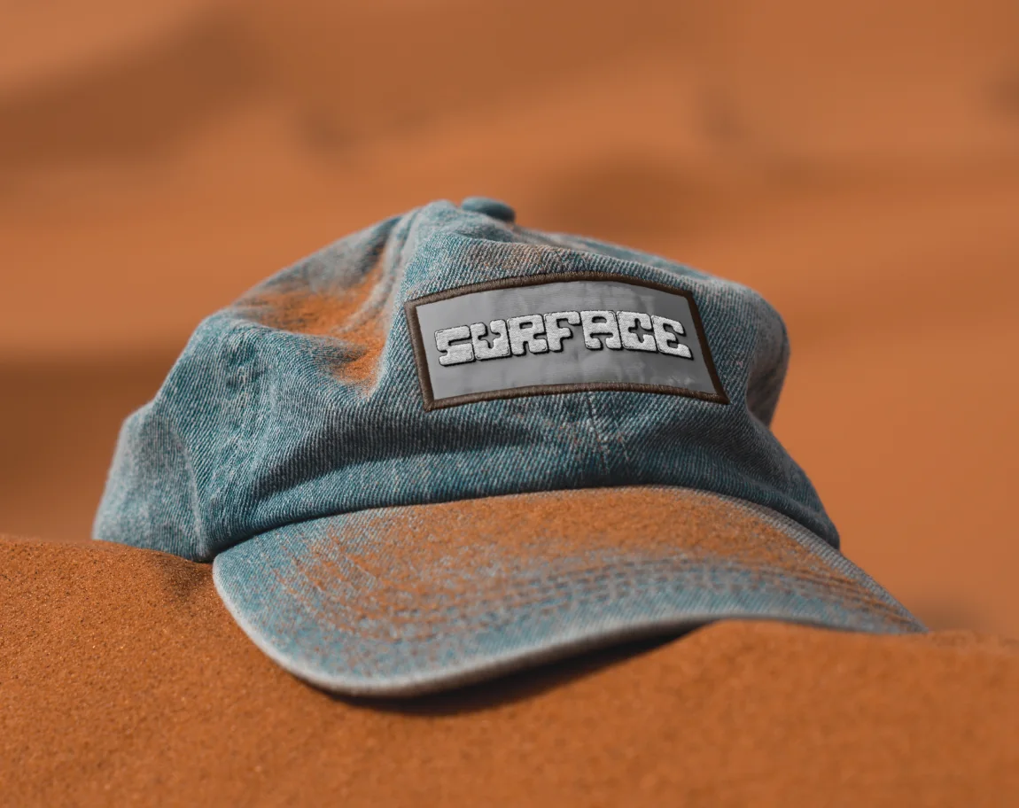 A washed denim cap with a Surface patch, on orange sand.